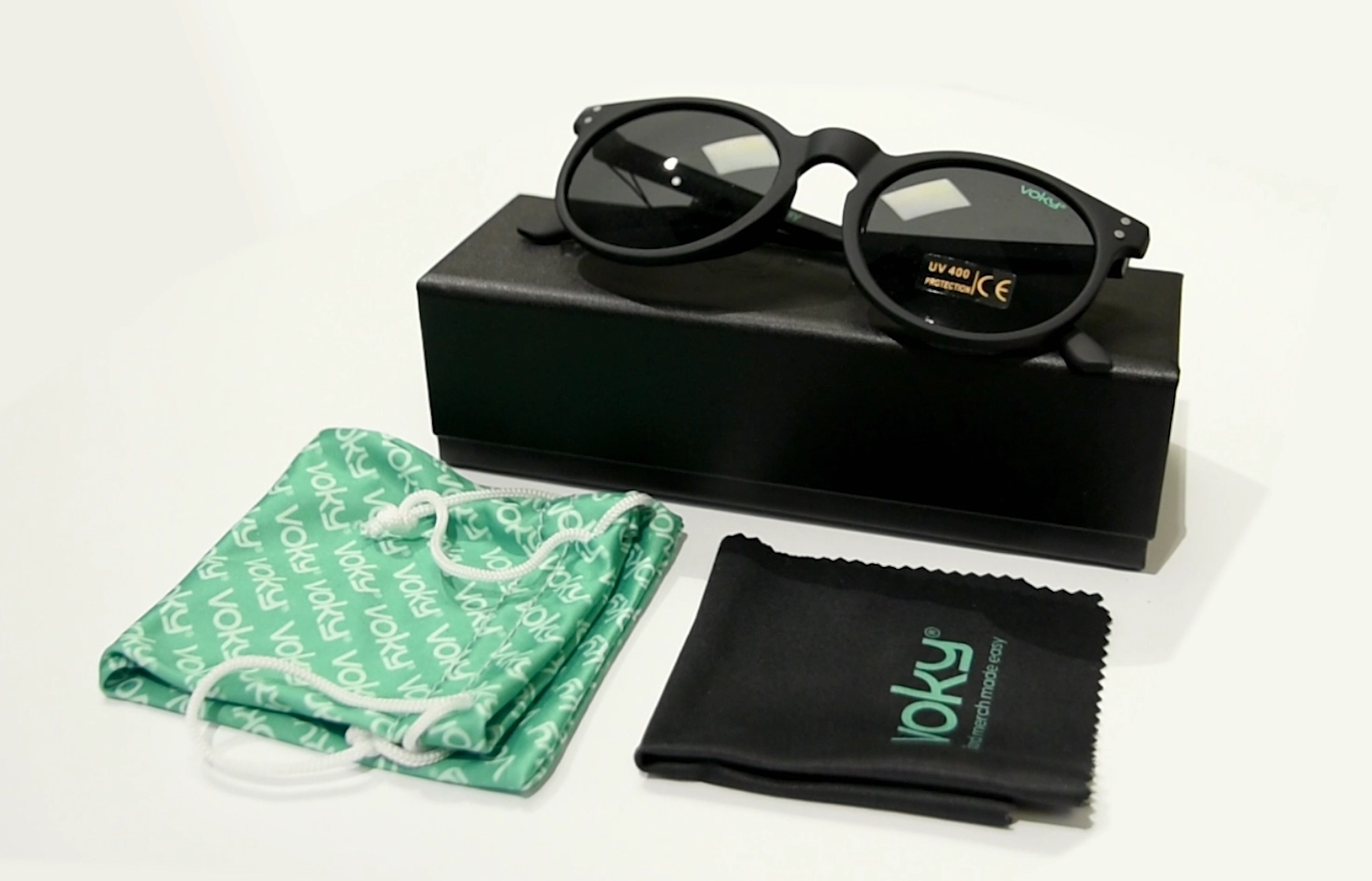 Fyra Voky-artiklar: lyxiga solglasögon med en liten logotyp på linsen, en svart låda, en grön mönstrad väska och en svart rengöringsduk med logotypen.
