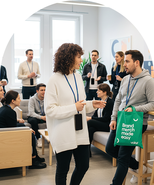 En kvinna i en vit tröja pratar med en man som håller i en grön tygkasse med texten "Brand merch made easy", omgiven av andra unga yrkesverksamma i en modern kontorsmiljö.